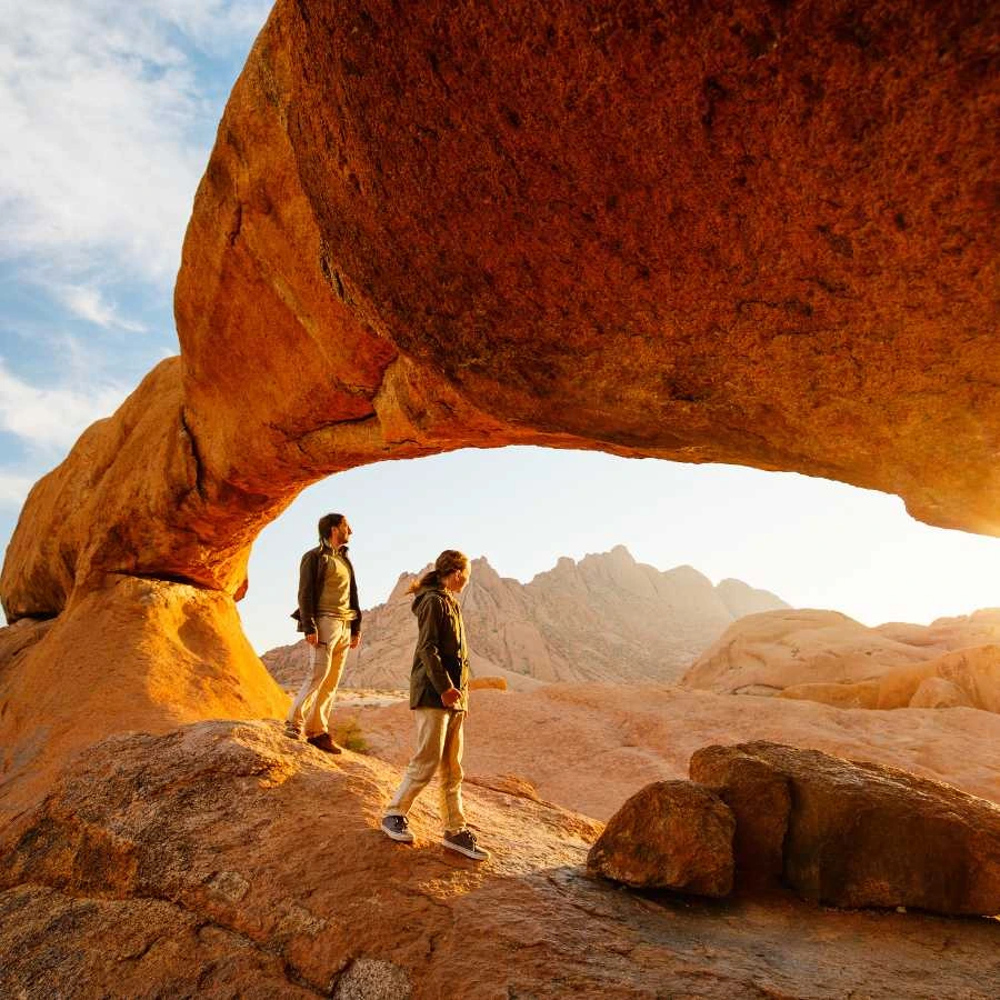 Viaggiatori esplorano una formazione rocciosa nel deserto della Namibia al tramonto</p>
<p>