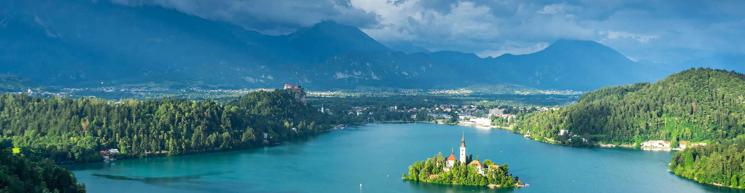 Veduta panoramica del Lago di Bled con isola, castello e montagne sullo sfondo, sotto un cielo nuvoloso