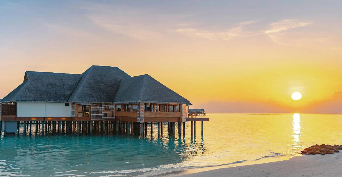 Resort di lusso su palafitta alle Maldive al tramonto, con vista sul mare calmo e cielo aranciato.