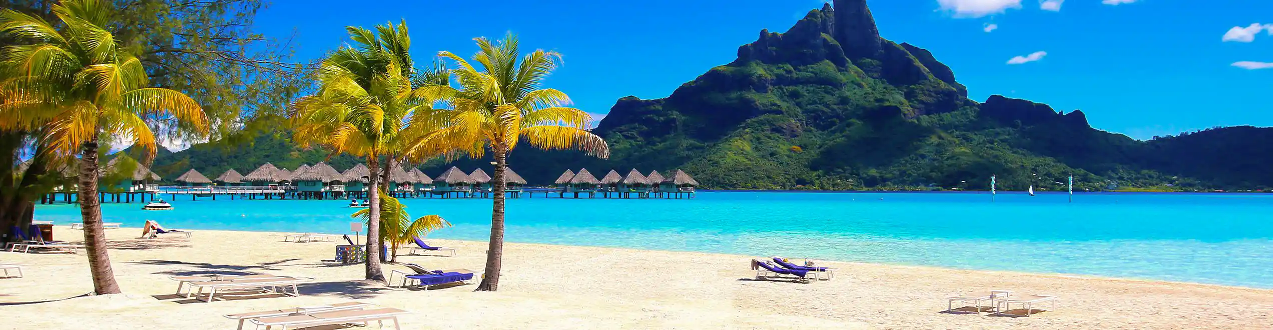 Spiaggia di Tahiti con sabbia bianca, palme e bungalow overwater davanti alla montagna verde.
