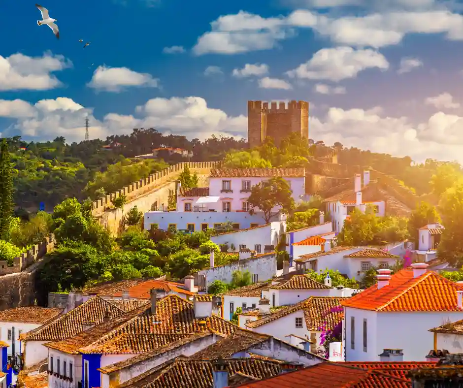 Óbidos incanta con le sue mura antiche, i tetti in terracotta e un’atmosfera sospesa nel tempo. Borgo medievale di Óbidos con case bianche, tetti rossi e castello sulle mura al tramonto