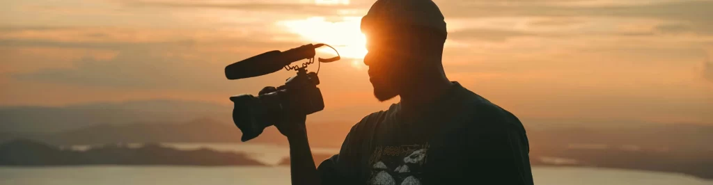 Videomaker controluce al tramonto con videocamera, su uno sfondo panoramico dal forte stile cinematografico