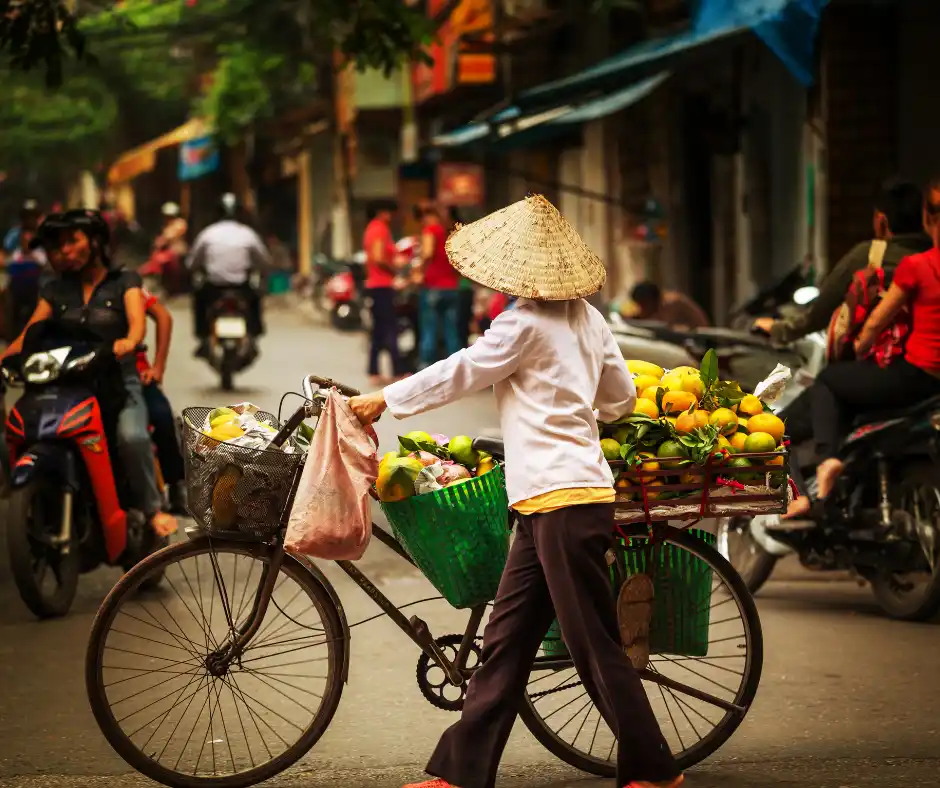 Offerta viaggio Vietnam tra cultura e vita locale autentica Didascalia Donna vietnamita con bicicletta carica di frutta tra le strade animate di Hanoi