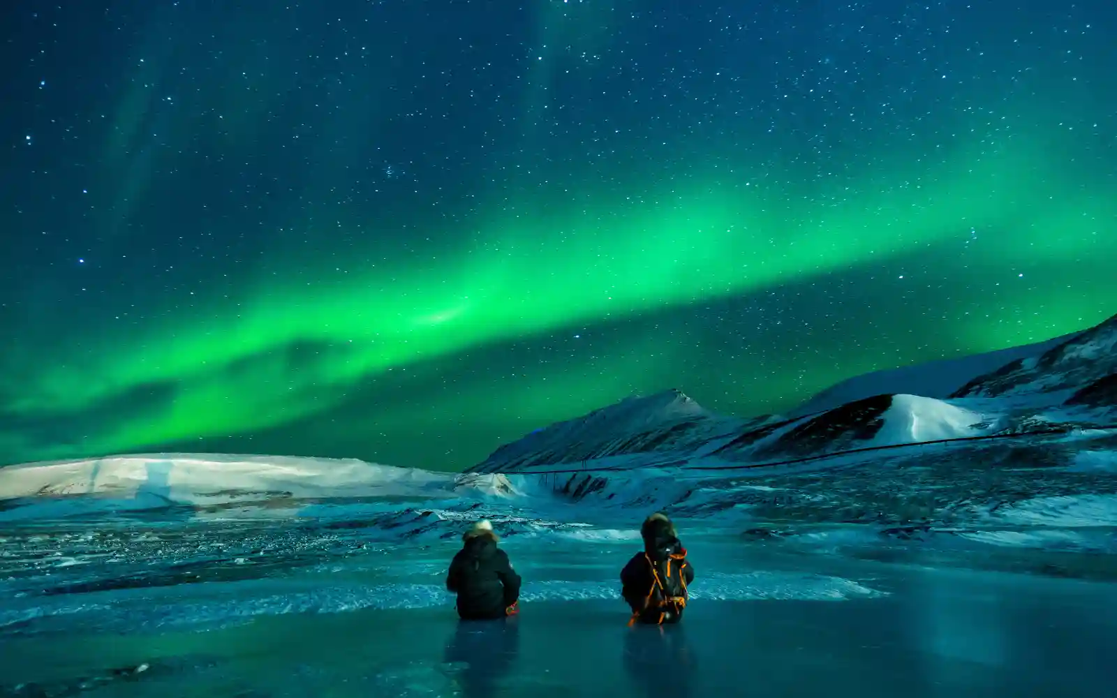 Due viaggiatori osservano l’aurora boreale davanti a montagne e distese di ghiaccio