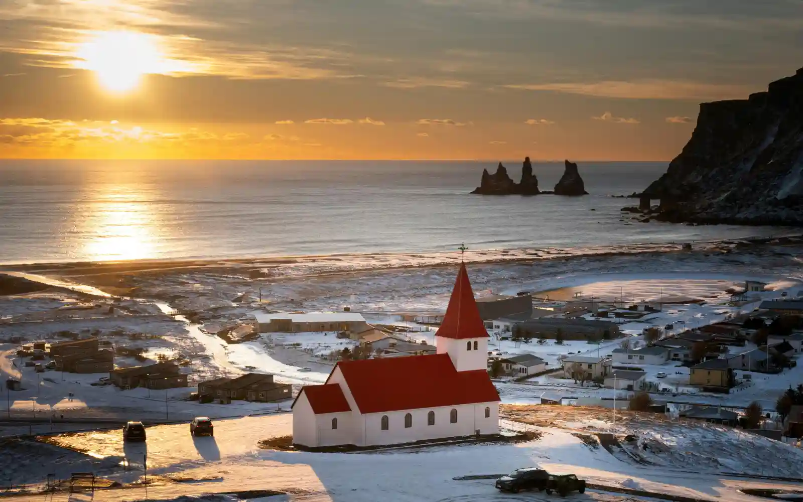Chiesa di Vík tra la neve con i faraglioni di Reynisdrangar sul mare