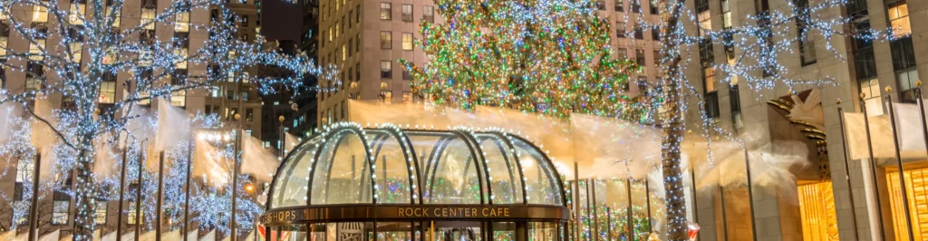 Albero di Natale illuminato al Rockefeller Center, dietro l’ingresso vetrato del Rock Center Cafe e alberi decorati con luci blu.