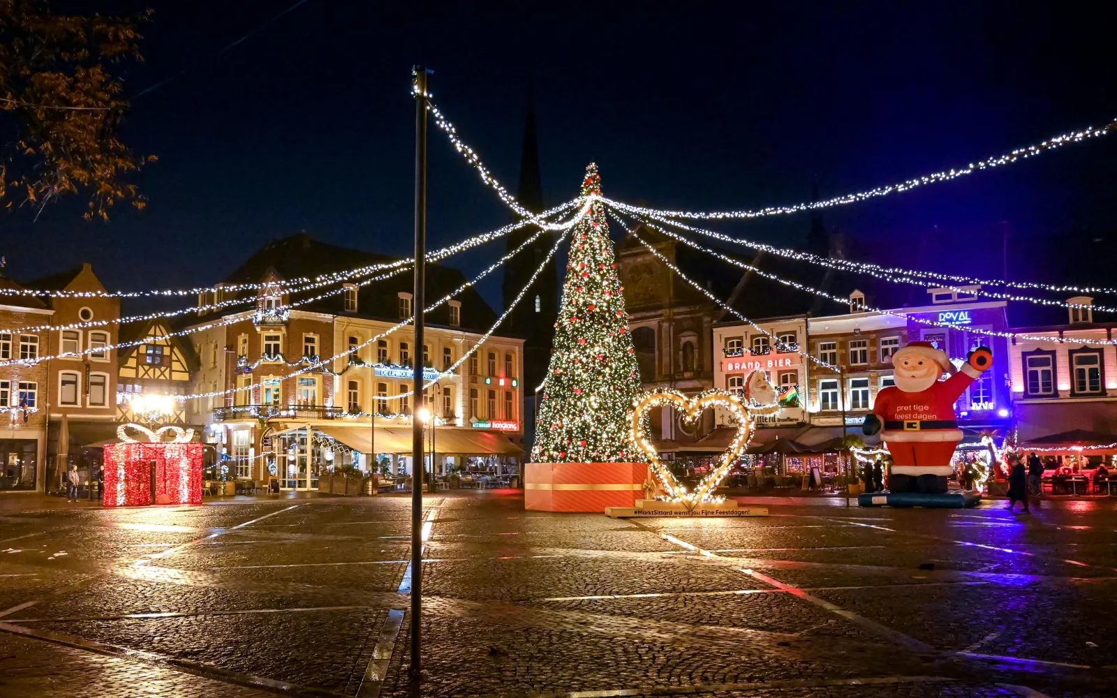 Grande albero di Natale illuminato in una piazza, con festoni luminosi e un Babbo Natale gonfiabile